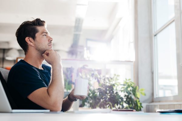 Side view shot of young businessman with coffee in hand sitting at his desk. Man sitting in office and thinking.