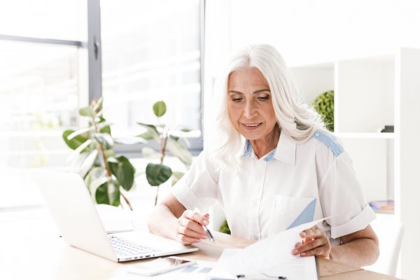 Picture of mature happy cheerful woman indoors in office working writing notes.