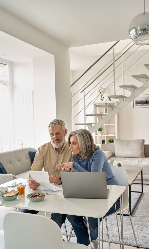 Senior mature couple check bank loan documents using laptop at home. Middle aged old family read paper bills payments, calculating pension or taxes, planning retirement finances, household expenses.